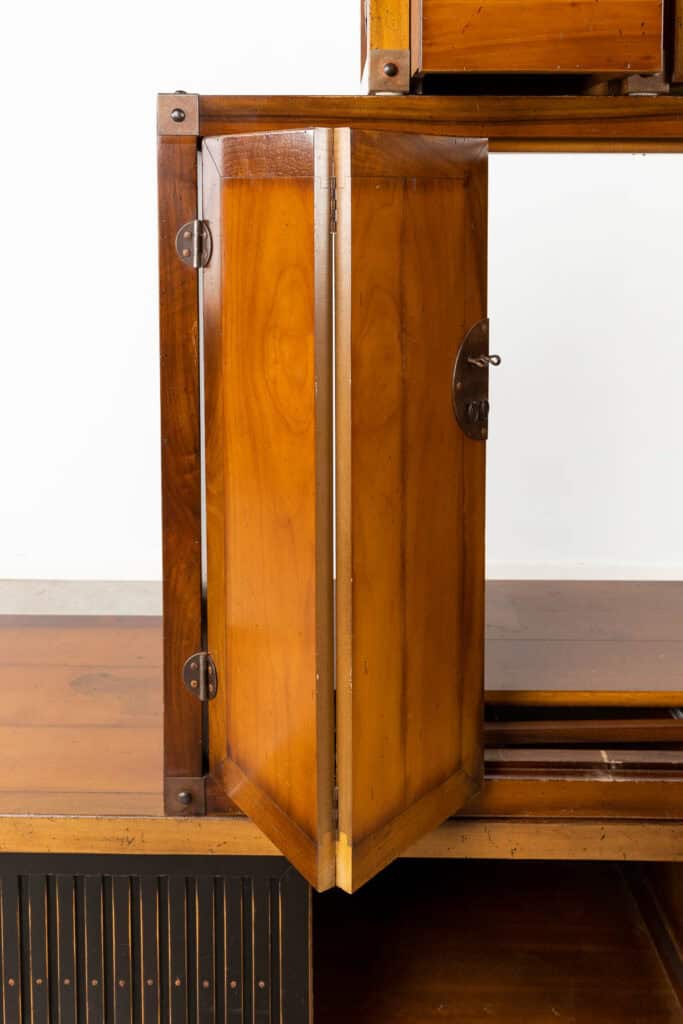 Een close-up van een houten kast met een gedeeltelijk open deur, waardoor het interieur zichtbaar is. De kast heeft metalen scharnieren en een ronde metalen handgreep. Het hout heeft een warme, gepolijste afwerking.