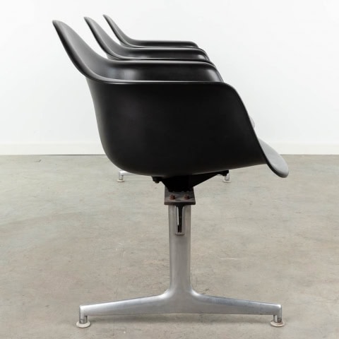 Een rij van vier aaneengeschakelde, zwarte plastic stoelen met metalen onderstellen, gezien vanaf de zijkant, tegen een eenvoudige witte muur en een betonnen vloer.