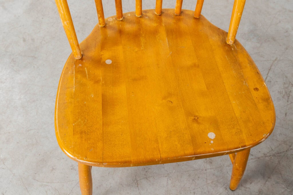 Een houten stoel met een gebogen zitting, met zichtbare krassen, slijtplekken en twee lichte verfvlekken op het oppervlak. De stoel staat op een grijze betonnen vloer.