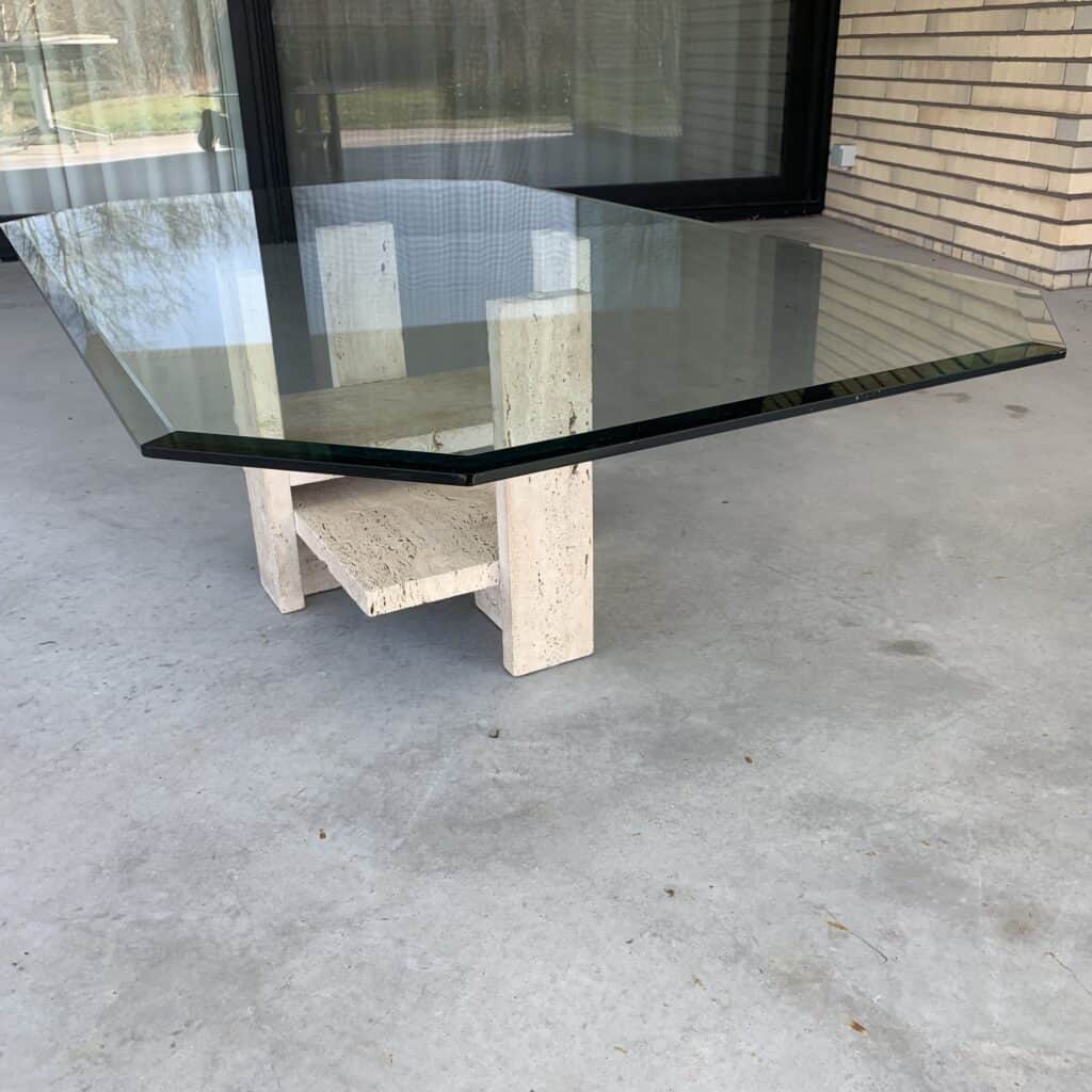 Een moderne salontafel met een dik, achthoekig glazen blad en een beige stenen onderstel staat op een betonnen terras buiten een gebouw met bakstenen muren en grote ramen.