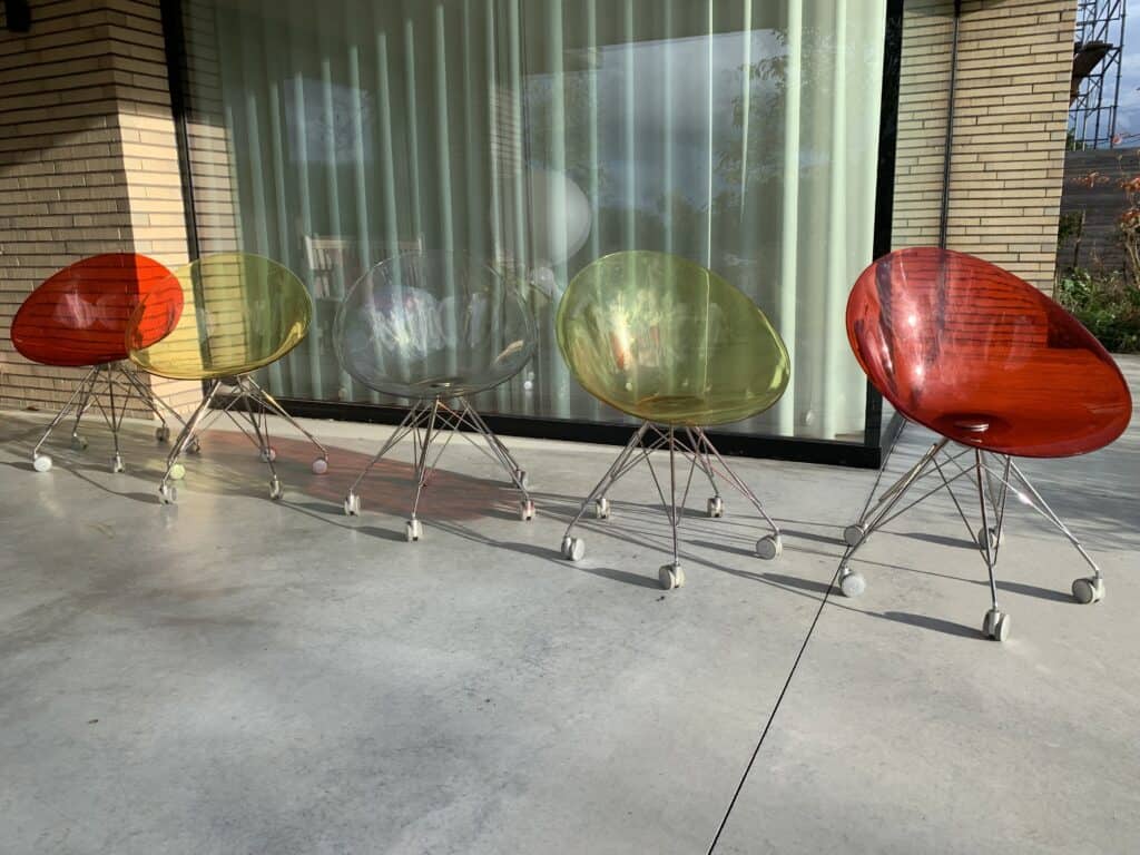 Vijf moderne, doorschijnende stoelen in de kleuren rood, geel en transparant staan op een rij op een betonnen terras voor een glazen raam met gordijnen. De stoelen hebben metalen poten en wieltjes.