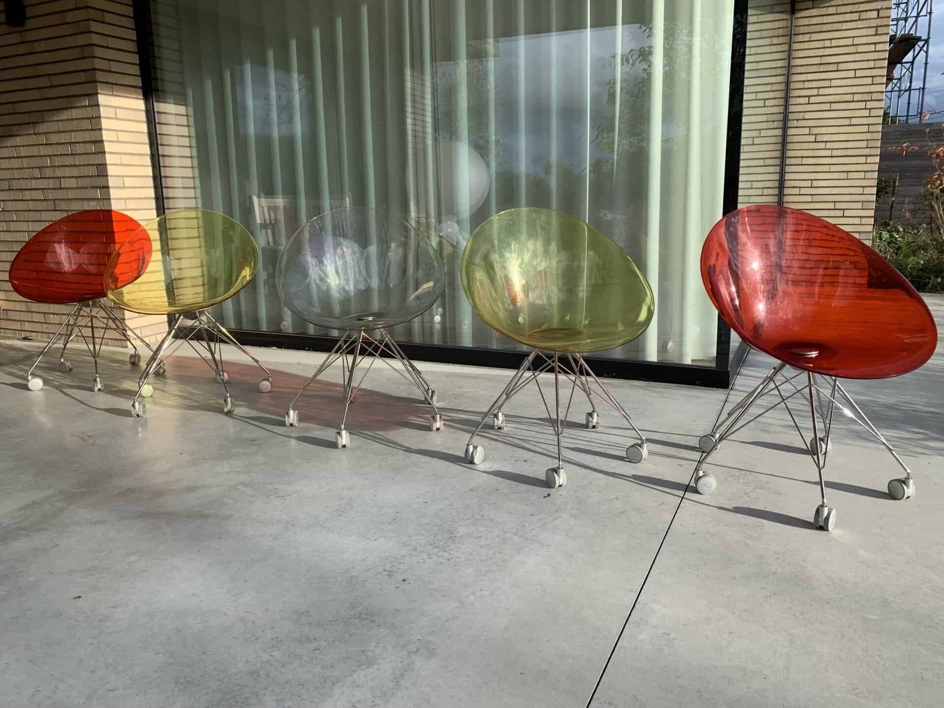 Vijf moderne, doorschijnende stoelen in de kleuren rood, geel en transparant staan op een rij op een betonnen terras voor een glazen raam met gordijnen. De stoelen hebben metalen poten en wieltjes.