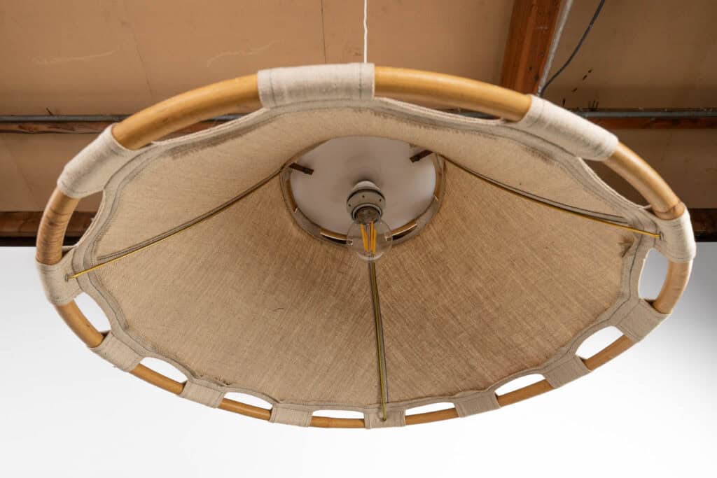 Een close-up van onderaf van een ronde lampenkap met een frame van bamboe en beige stof gespannen tussen de steunen, hangend aan een plafond met zichtbare balken.