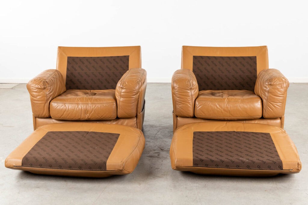 Twee beige leren fauteuils met bruine stoffen kussens en bijpassende voetenbankjes. De stof heeft een herhalend patroon en beide stoelen staan op een grijze vloer tegen een effen witte achtergrond.