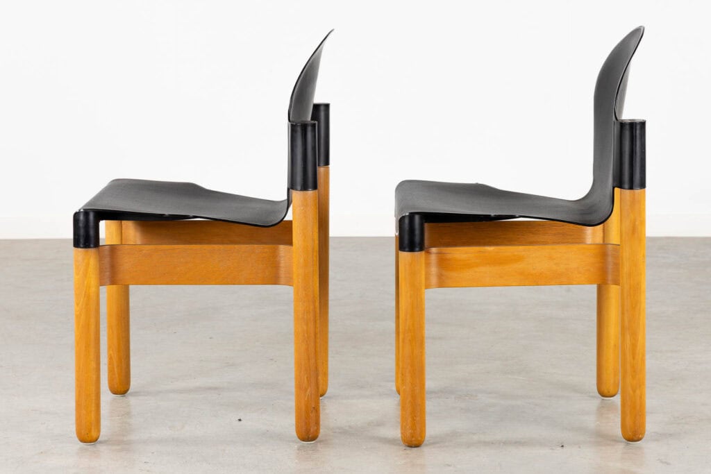 Twee moderne stoelen met gebogen, zwarte zittingen en rugleuningen en stevige houten frames staan naast elkaar, beide naar links gericht, tegen een effen witte muur en een betonnen vloer als achtergrond.