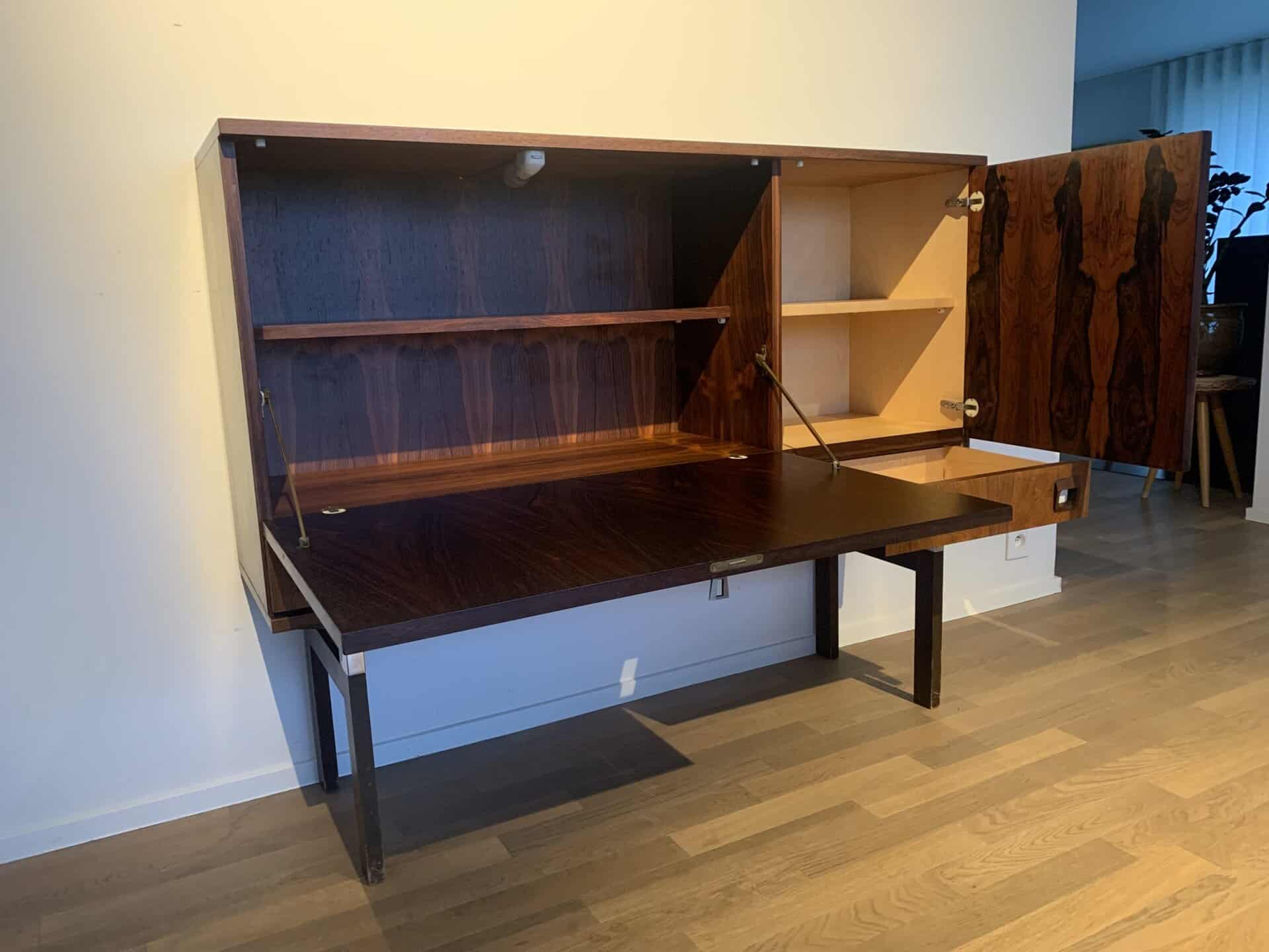 Een uitklapbaar houten bureau voor aan de muur met open kasten en planken, waardoor opbergruimte en een ingebouwd werkblad zichtbaar zijn. Het meubel is bevestigd aan een lichtgekleurde muur boven een houten vloer.