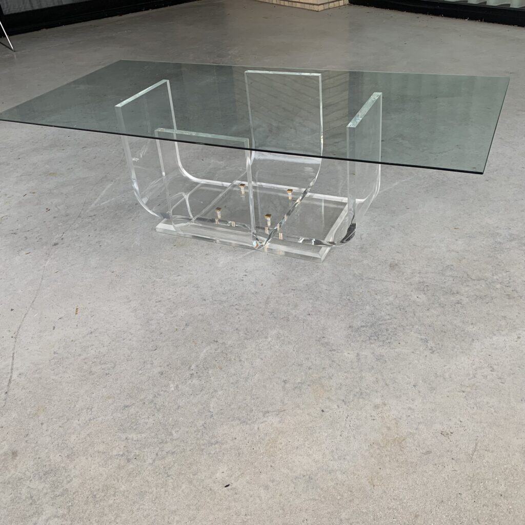 Een moderne glazen tafel met een rechthoekig blad van helder glas en een geometrisch, transparant onderstel van acryl, op een lichtgrijze betonnen vloer.