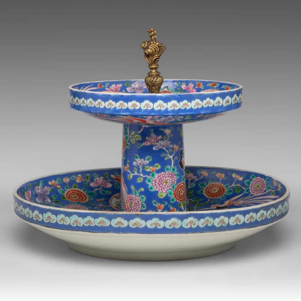 Een porseleinen onderbord met twee niveaus, versierd met ingewikkelde bloemen- en vogelmotieven op een levendige blauwe achtergrond, bekroond met een sierlijke gouden knop. De onder- en bovenborden zijn rond met decoratieve randen.