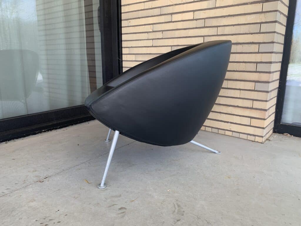 Een moderne, zwarte stoel met een rond, hoekig ontwerp en dunne metalen poten staat op een betonnen terras bij een glazen deur en een lichte bakstenen muur.