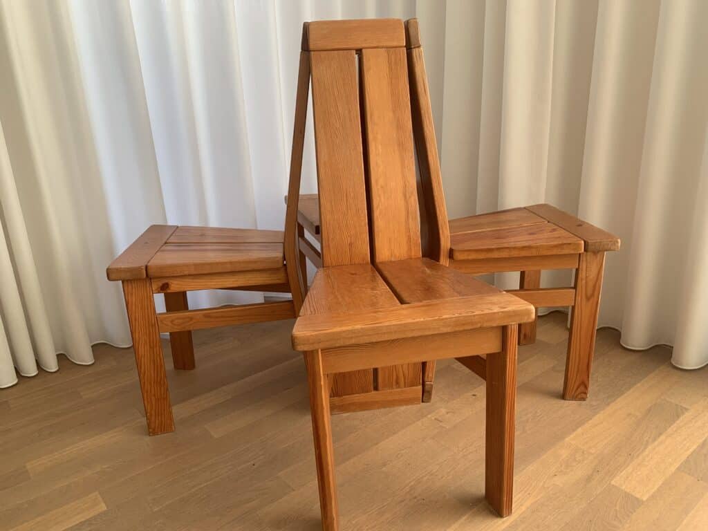 Drie houten stoelen staan in een driehoekige opstelling, met de rugleuningen in elkaar grijpend, op een lichte houten vloer en met witte gordijnen op de achtergrond.