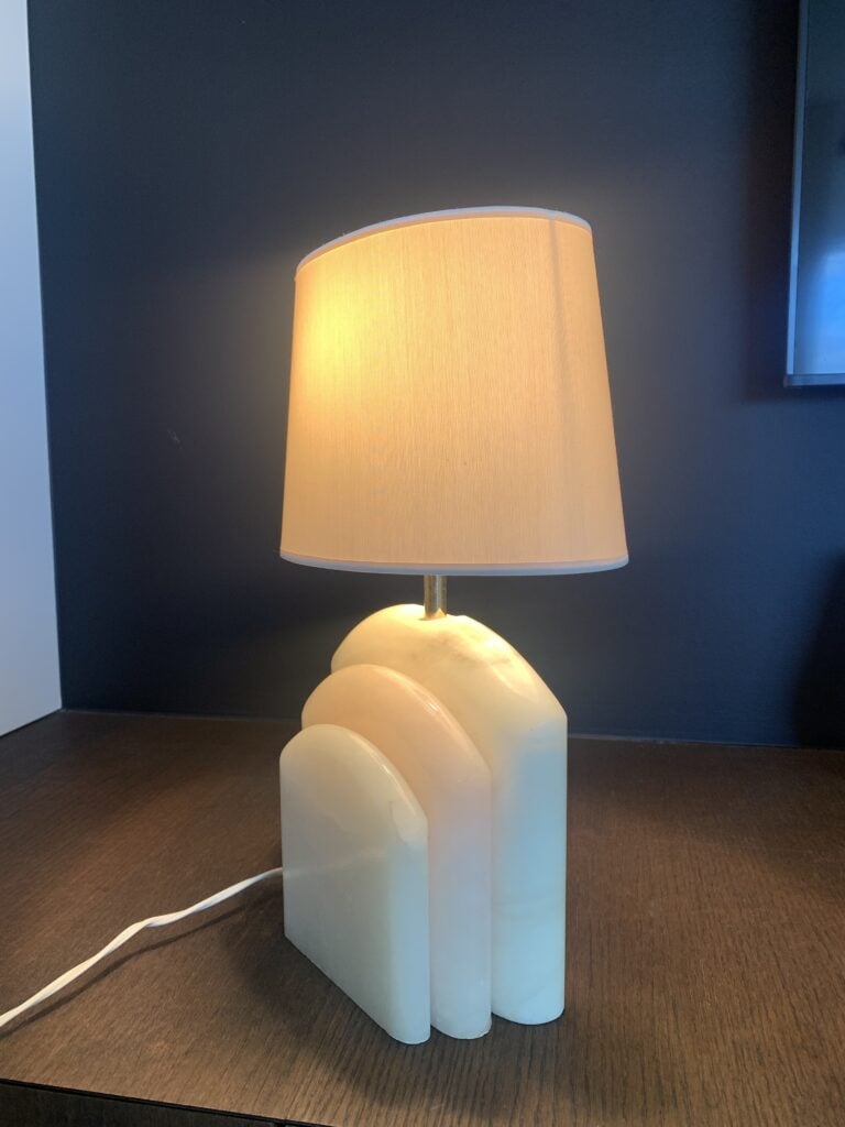 Een tafellamp met een gebogen, gelaagde witte voet en een beige stoffen lampenkap staat op een donker houten oppervlak tegen een donkere achtergrond. De lamp is aan en werpt een warm licht.