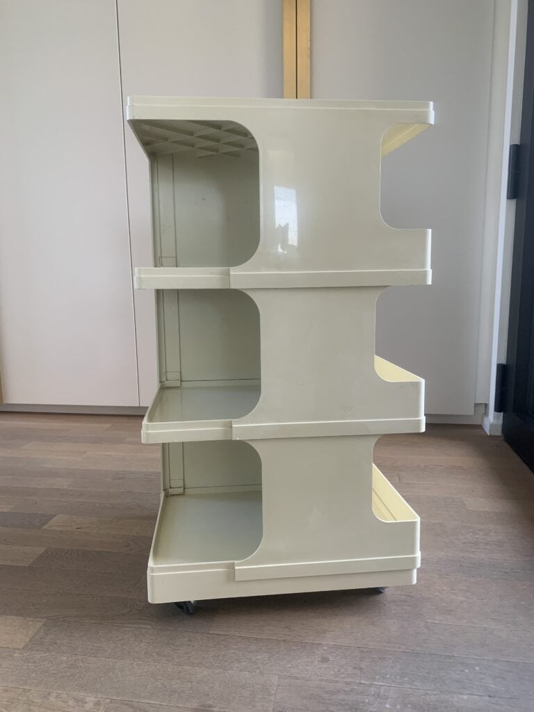 Een beige, drielaags, kunststof opbergkarretje met open planken en wielen staat op een houten vloer in een moderne kamer.