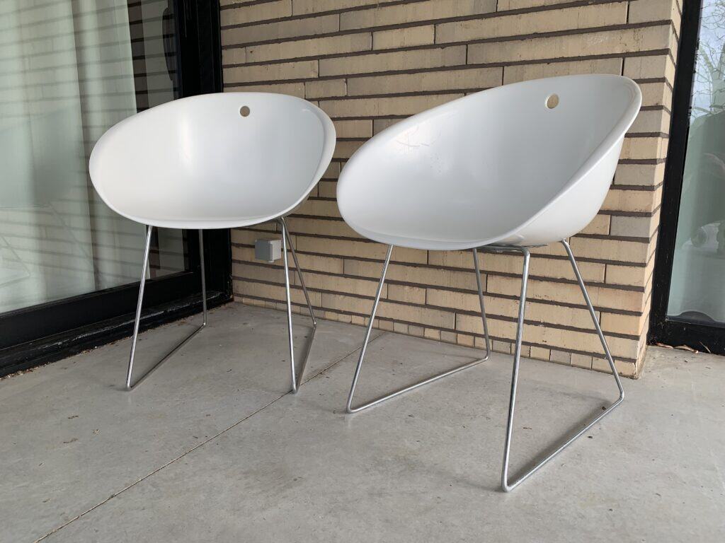 Twee moderne witte stoelen met metalen poten staan op een betonnen terras voor een bakstenen muur en grote glazen deuren.