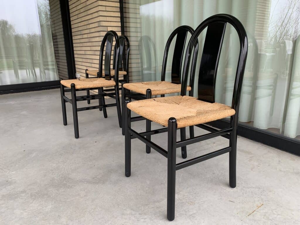 Drie zwarte stoelen met gevlochten rieten zittingen staan op een rij op een betonnen terras, voor grote ramen met doorschijnende witte gordijnen en een bakstenen muur. Buiten zijn bomen en een gazon te zien.