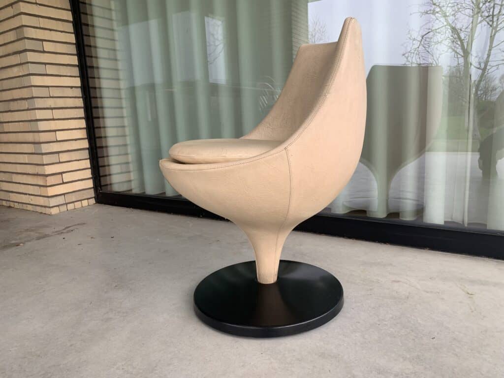 Een moderne beige draaistoel met een ronde zitting en rugleuning, voorzien van een enkele zwarte voet, staat op een betonnen vloer voor een glazen deur met witte gordijnen.