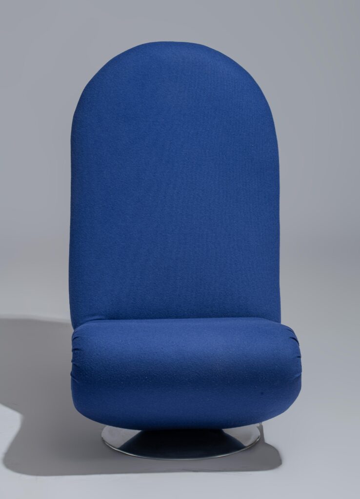 Een moderne stoel met hoge rugleuning, bekleed met blauwe stof, een afgeronde bovenkant en een rond metalen onderstel, tegen een effen grijze achtergrond.