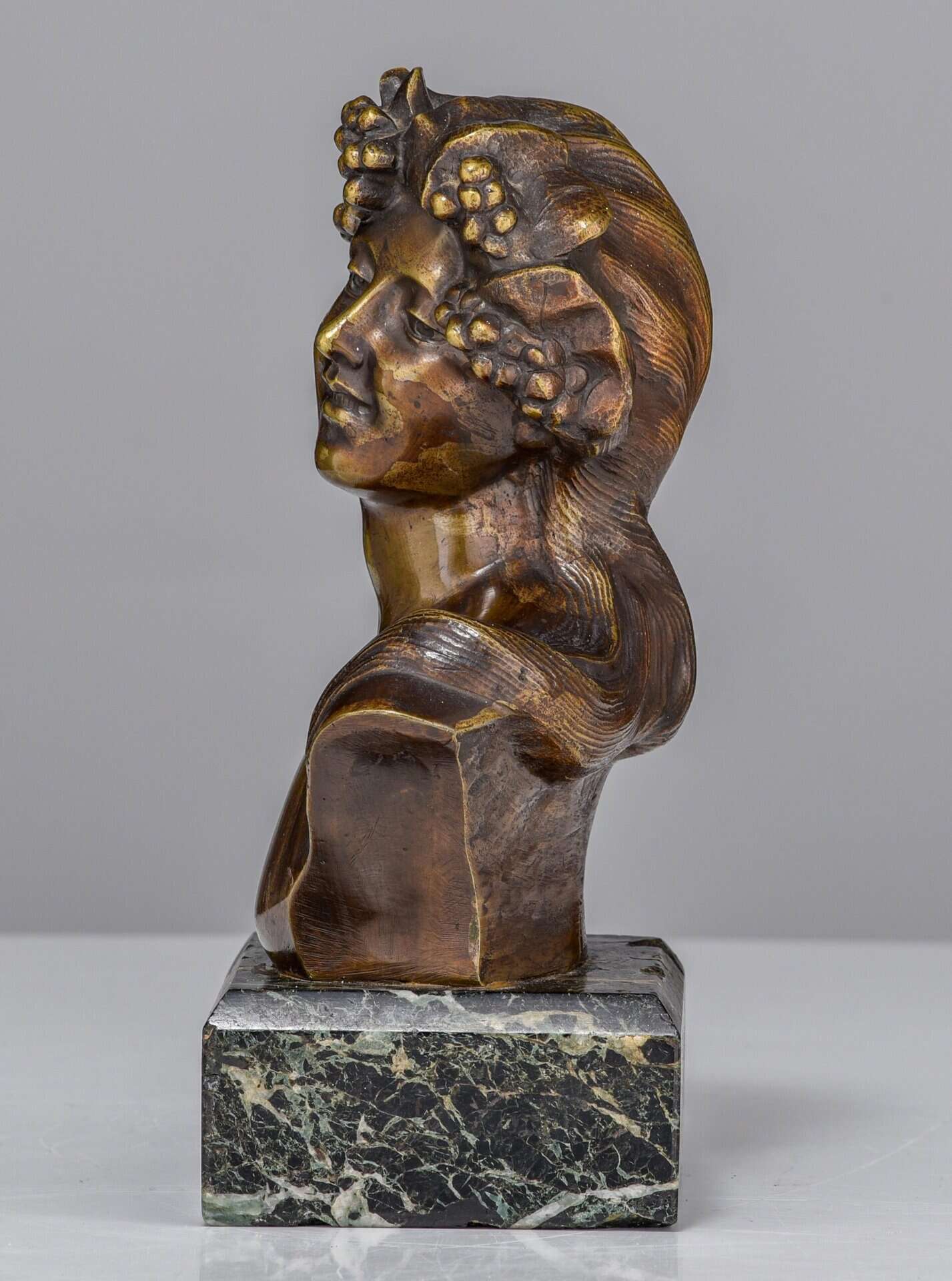 Bronzen buste van een vrouw met druivenbladeren en -trossen in haar haar, geplaatst op een vierkante zwarte marmeren sokkel. De figuur kijkt omhoog met een serene uitdrukking.