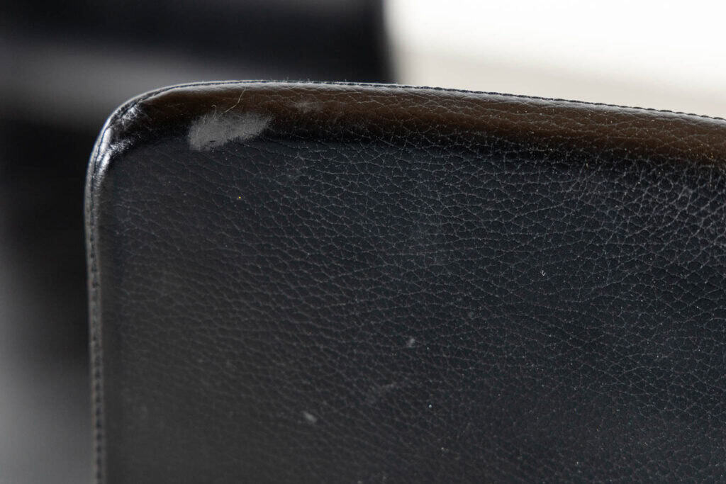 Close-up van de rand van een zwarte leren stoel met een zichtbare slijtplek bij de hoek. Hierop is lichte slijtage te zien en de textuur van het leer is goed te zien.