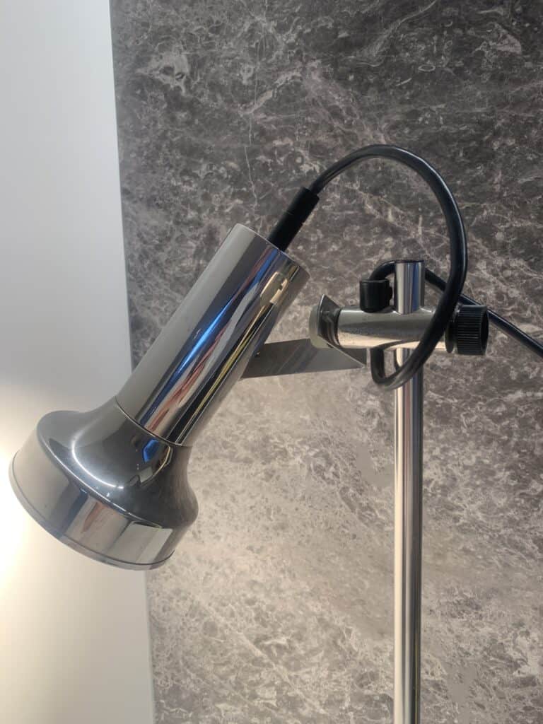 Een close-up van een moderne, metalen bureaulamp met een buisvormig ontwerp, bevestigd aan een verticale standaard voor een getextureerde grijs-witte muur. Het zwarte netsnoer loopt over de lamp.