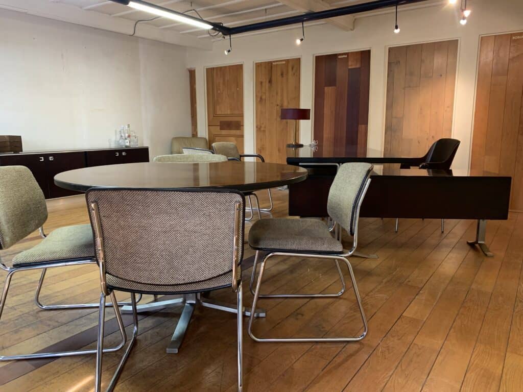 Een modern kantoor met een houten vloer, een ronde tafel met vier stoelen, een bureau met een stoel en een lamp, en houten wandpanelen op de achtergrond. De kamer is goed verlicht met spots.