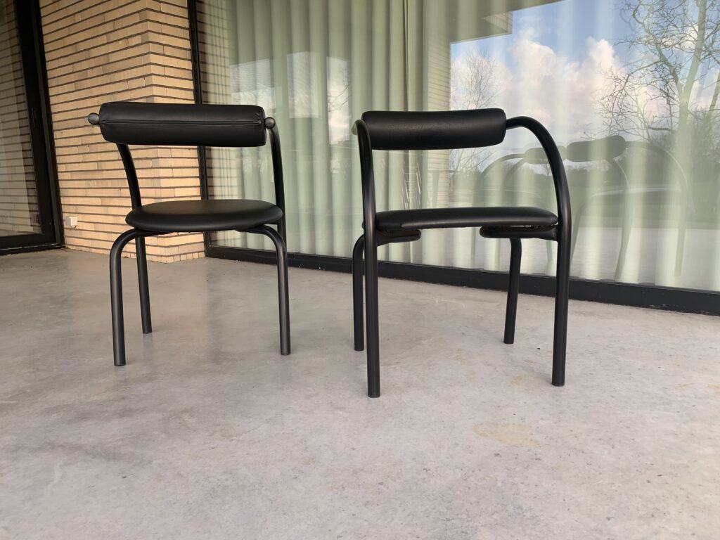 Twee moderne zwarte stoelen met gebogen rugleuningen en poten staan op een glad betonnen terras. Achter de stoelen bevindt zich een groot raam met gesloten gordijnen en een bakstenen muur aan de linkerkant.