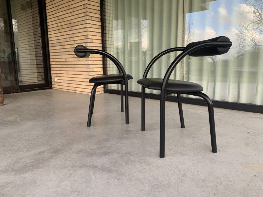 Twee moderne zwarte stoelen met gebogen rugleuningen staan op een betonnen terras voor een gebouw met bakstenen muren en grote glazen ramen, afgedekt met witte gordijnen. Bomen en de lucht worden in het glas weerspiegeld.