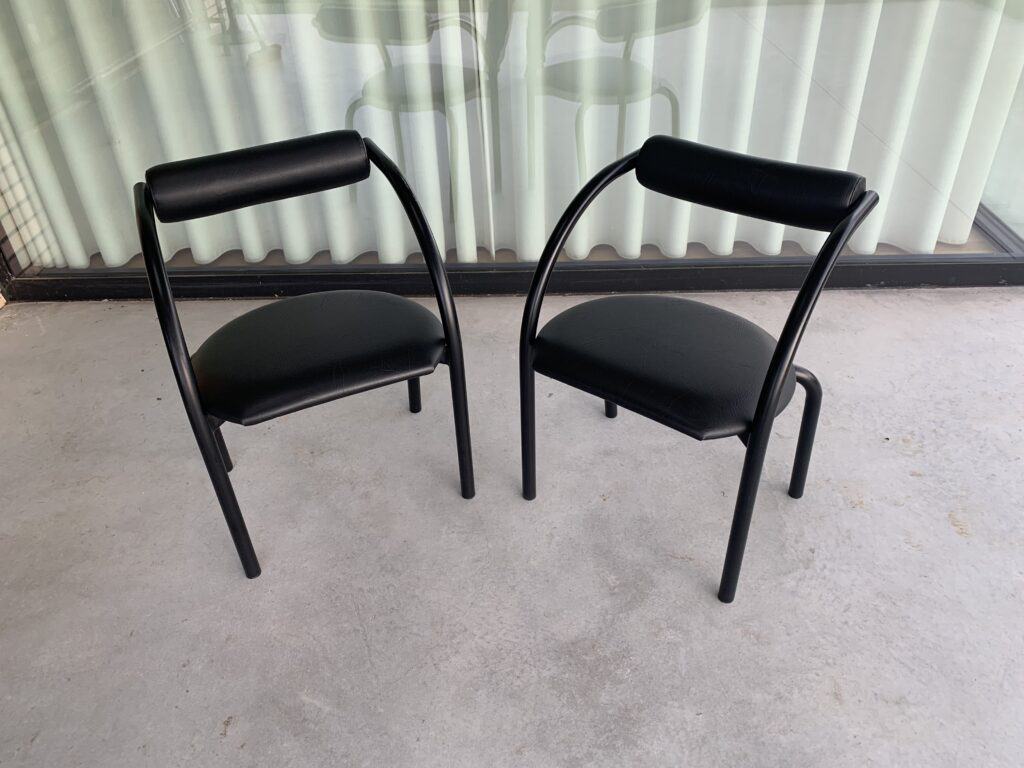 Twee moderne zwarte stoelen met gebogen poten en rugleuningen staan op een betonnen vloer voor een raam met verticale jaloezieën. Het ontwerp is minimalistisch en eigentijds.