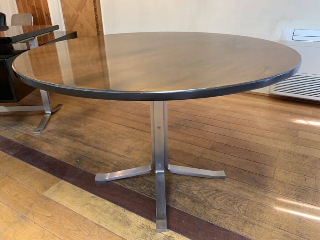 Een ronde houten tafel met een metalen voetstuk staat op een hardhouten vloer in een kamer met beige muren en zichtbaar meubilair op de achtergrond.