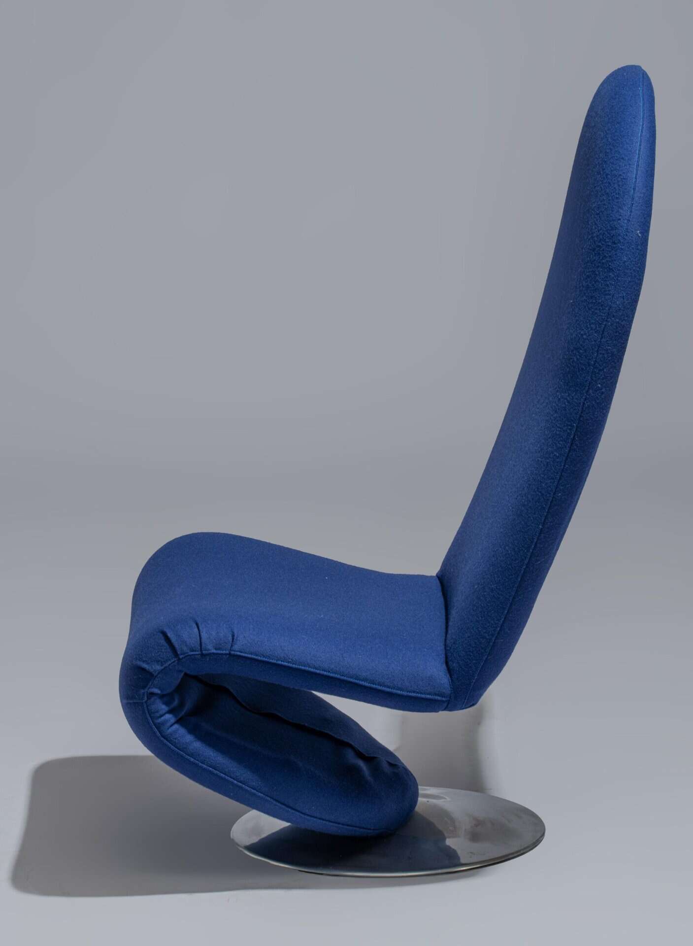 Een moderne blauwe loungestoel met een gebogen, sculpturale vorm en een hoge rugleuning, geplaatst op een ronde metalen basis tegen een effen grijze achtergrond.