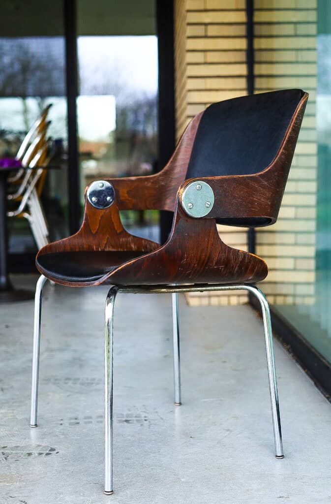 Een moderne stoel met een gebogen zitting van donker hout, een zwarte rugleuning en metalen poten staat op een terras voor glazen deuren en een bakstenen muur. Op de achtergrond staan nog meer stoelen opgestapeld.