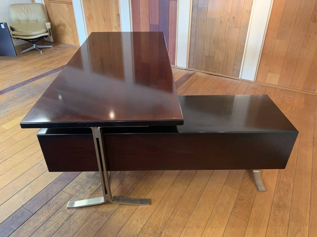 Een modern, donkerhouten directiebureau met een strak, schuin design en metalen poten, staat op een lichte houten vloer met houten lambrisering op de achtergrond. In de hoek is een bureaustoel zichtbaar.