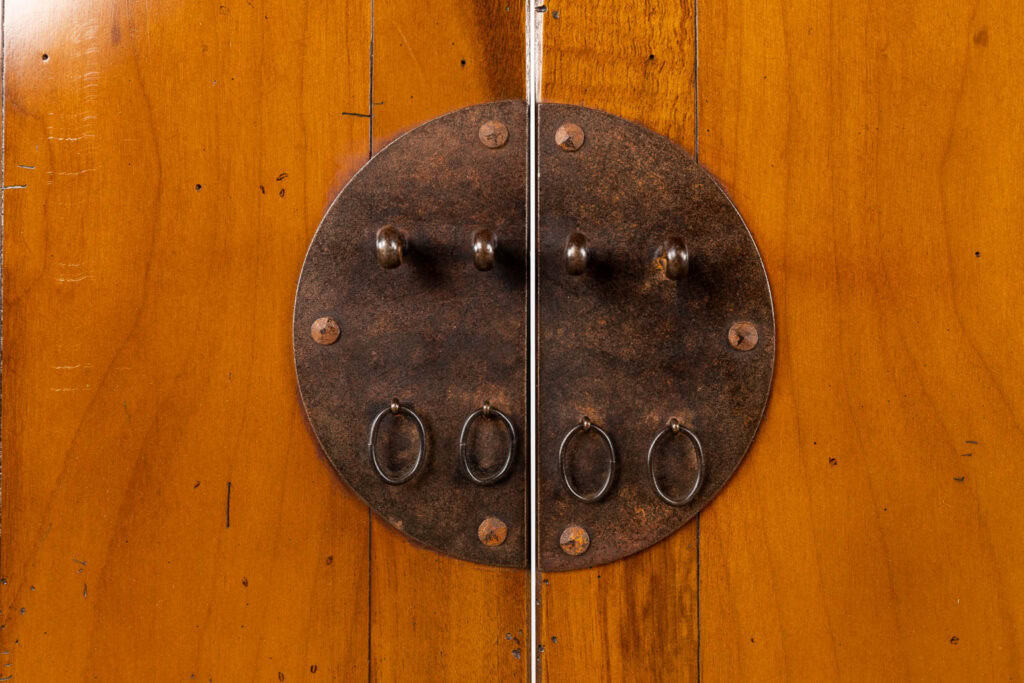 Close-up van een houten kast met een ronde, rustieke metalen sluitplaat en vier kleine metalen ringen, twee aan elke kant waar de kastdeuren in het midden samenkomen.