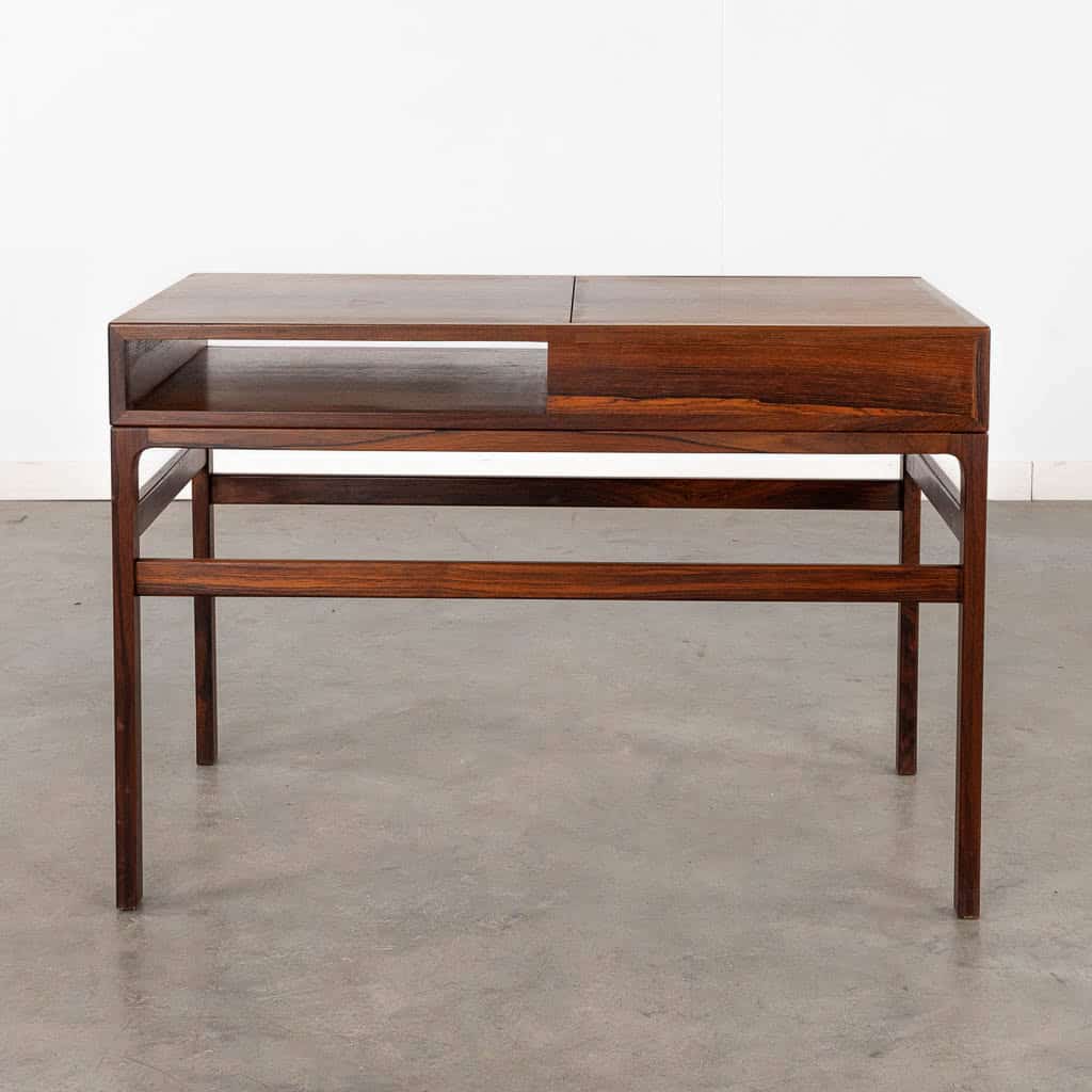 Een minimalistisch houten bureau met strakke lijnen, open opbergruimte aan de linkerkant en een gesloten vak aan de rechterkant, staand op vier slanke poten op een grijze betonnen vloer tegen een eenvoudige witte muur.