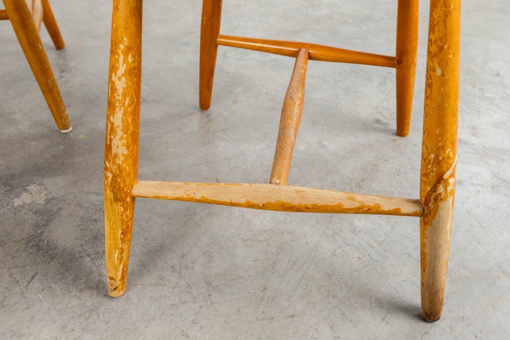 Close-up van de poten van een houten stoel met versleten en afgebladerde vernis, waardoor kale plekken zichtbaar zijn. De stoel staat op een grijze betonnen vloer.