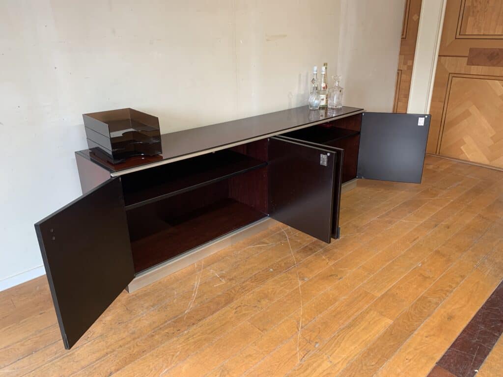 Een lang, donkerhouten dressoir met twee open kastdeuren, met lege planken erin. Bovenop staan wat flessen en een zwarte organizer. Het meubel staat op een houten vloer tegen een effen muur.