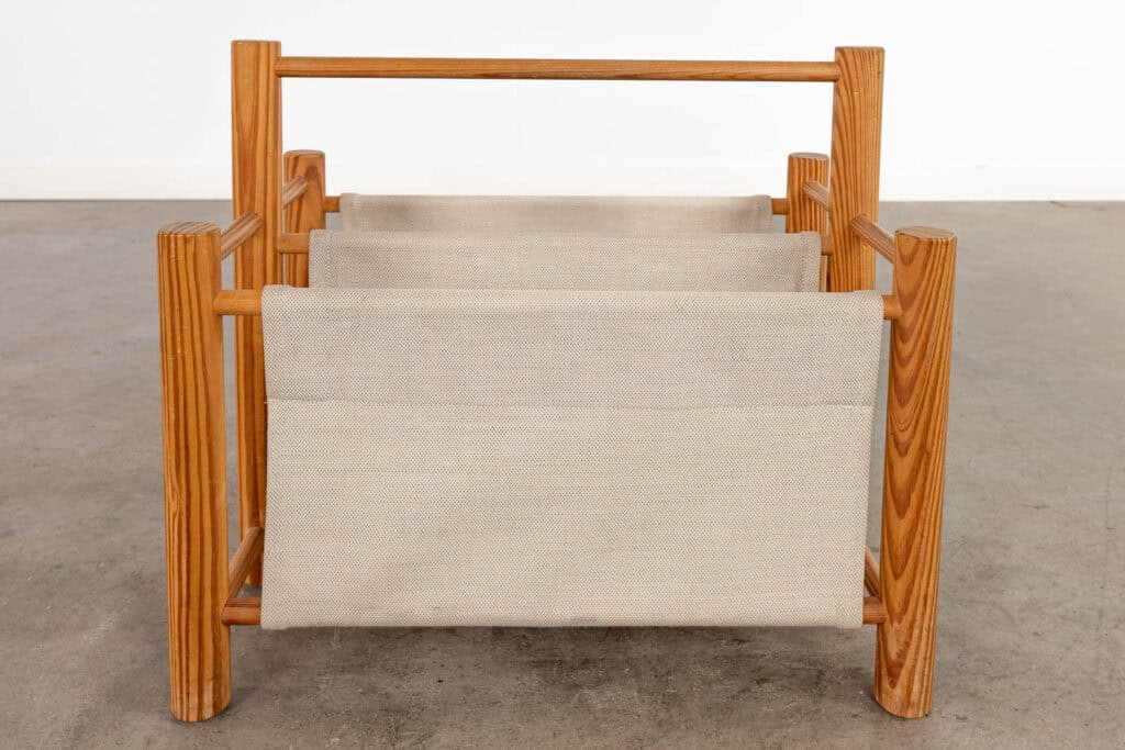 Een houten tijdschriftenrek met drie beige canvas draagbanden, geplaatst op een grijze betonnen vloer tegen een witte muur.