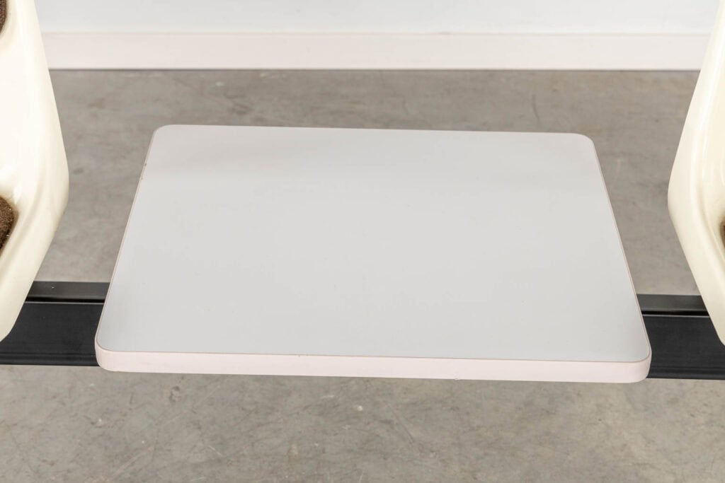 Tussen twee crèmekleurige stoelen staat een rechthoekig, wit tafelblad of dienblad. Daaronder is de grijze, betonnen vloer zichtbaar.