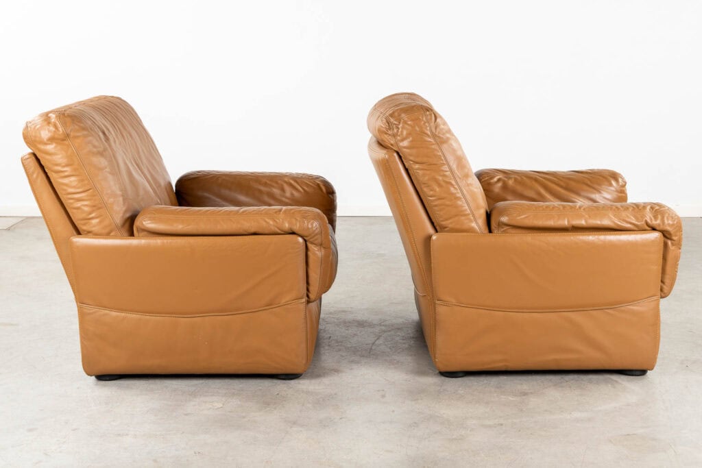 Twee bruine leren fauteuils staan naast elkaar, in tegenovergestelde richting, op een lichtgrijze vloer met een effen witte achtergrond.