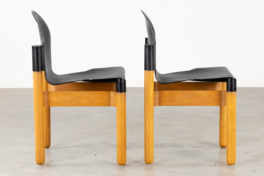 Twee moderne stoelen met zwarte kunststof zittingen en rugleuningen en lichte houten poten en frames staan naast elkaar, in tegenovergestelde richting, op een effen betonnen vloer tegen een witte achtergrond.