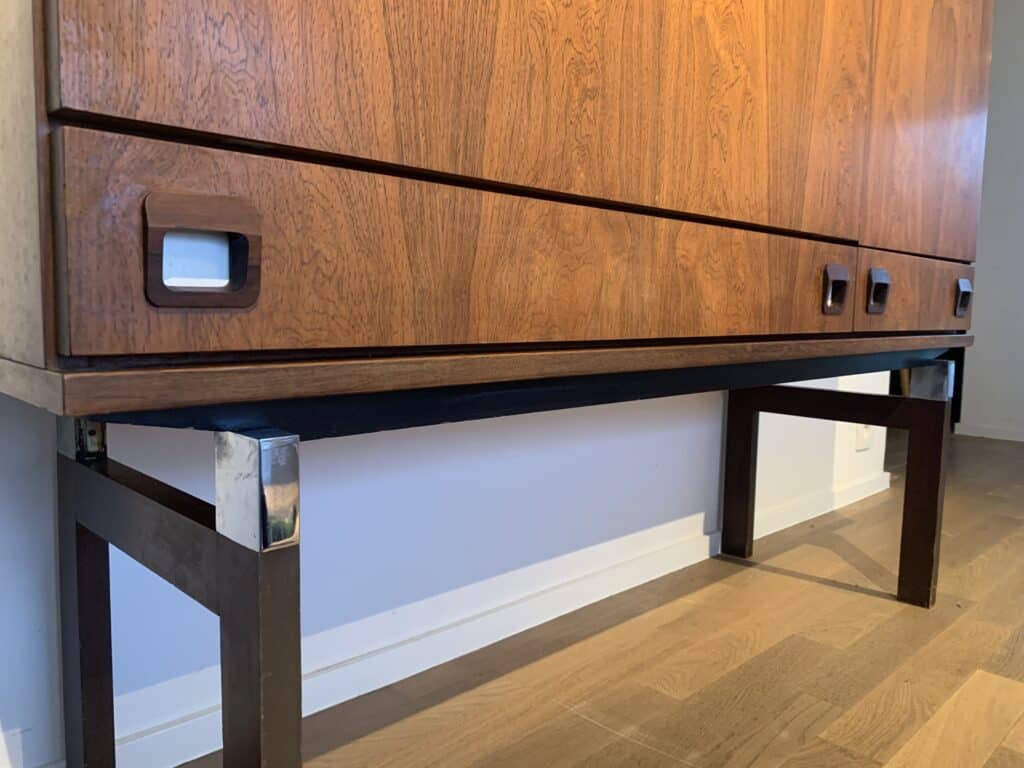 Een close-up van een houten kast in mid-century modern-stijl met schuifdeuren en donkere rechthoekige poten, geplaatst tegen een witte muur op een houten vloer.