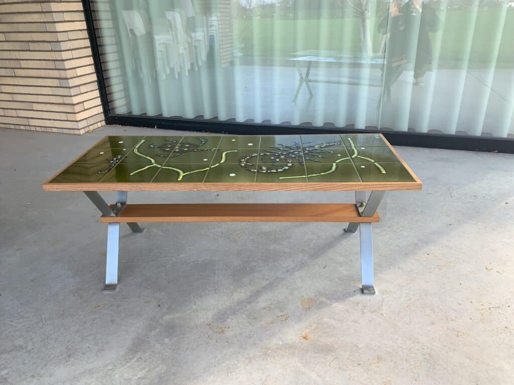 Een rechthoekige tafel met een groen, decoratief tegelblad en metalen poten staat op een betonnen terras. Op de achtergrond zijn een glazen wand en een bakstenen muur te zien, met daarachter terrasstoelen.