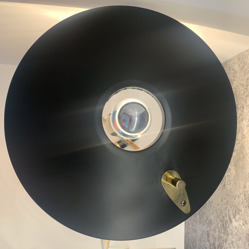 Een close-up van een moderne lamp met een grote, zwarte, ronde kap, een reflecterend zilveren middenstuk en een kleine, gouden verstelhendel aan de zijkant. Het licht schijnt recht in de camera.