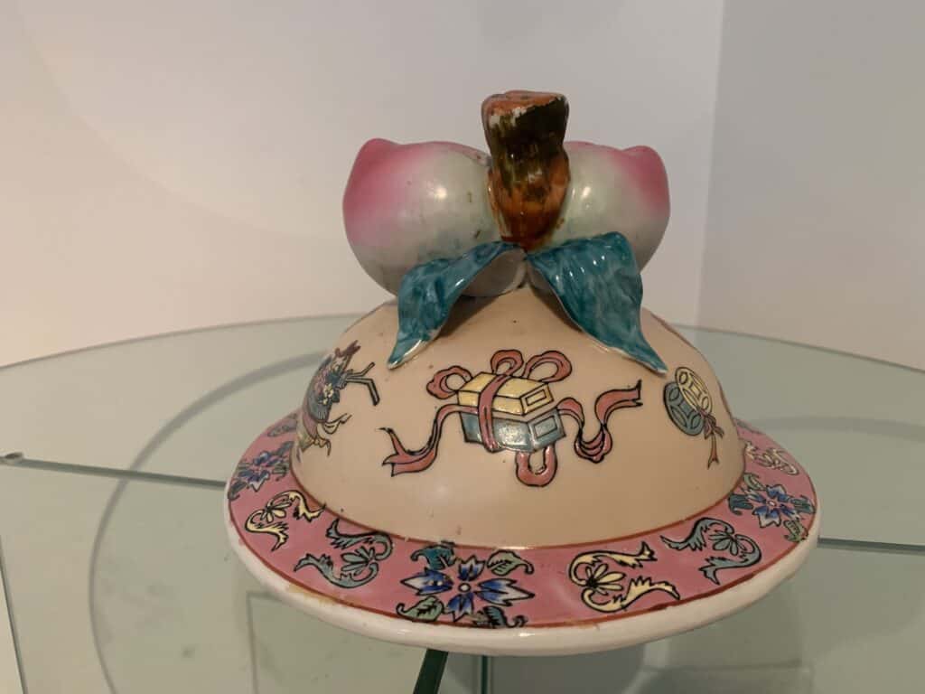 Een decoratieve keramische schaal met deksel, versierd met pastelroze en blauwe dessins. Het deksel is beschilderd met symbolen en is versierd met een gebeeldhouwde perzik en bladeren als handvat. De schaal staat op een glazen oppervlak.