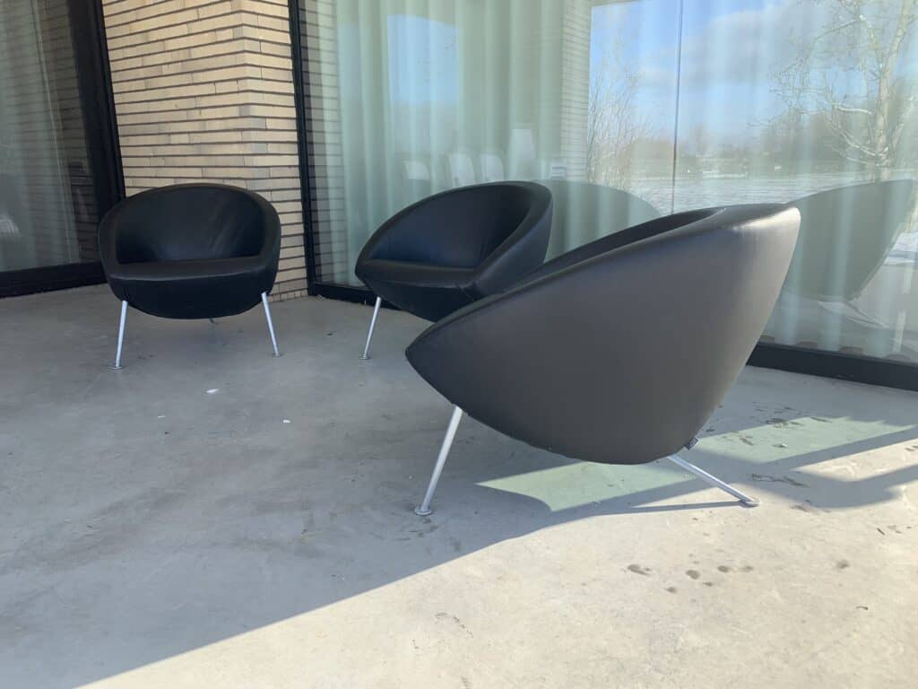Drie moderne zwarte stoelen met metalen poten staan op een betonnen terras bij grote glazen ramen met witte gordijnen. Het zonlicht werpt schaduwen en links is een bakstenen muur.