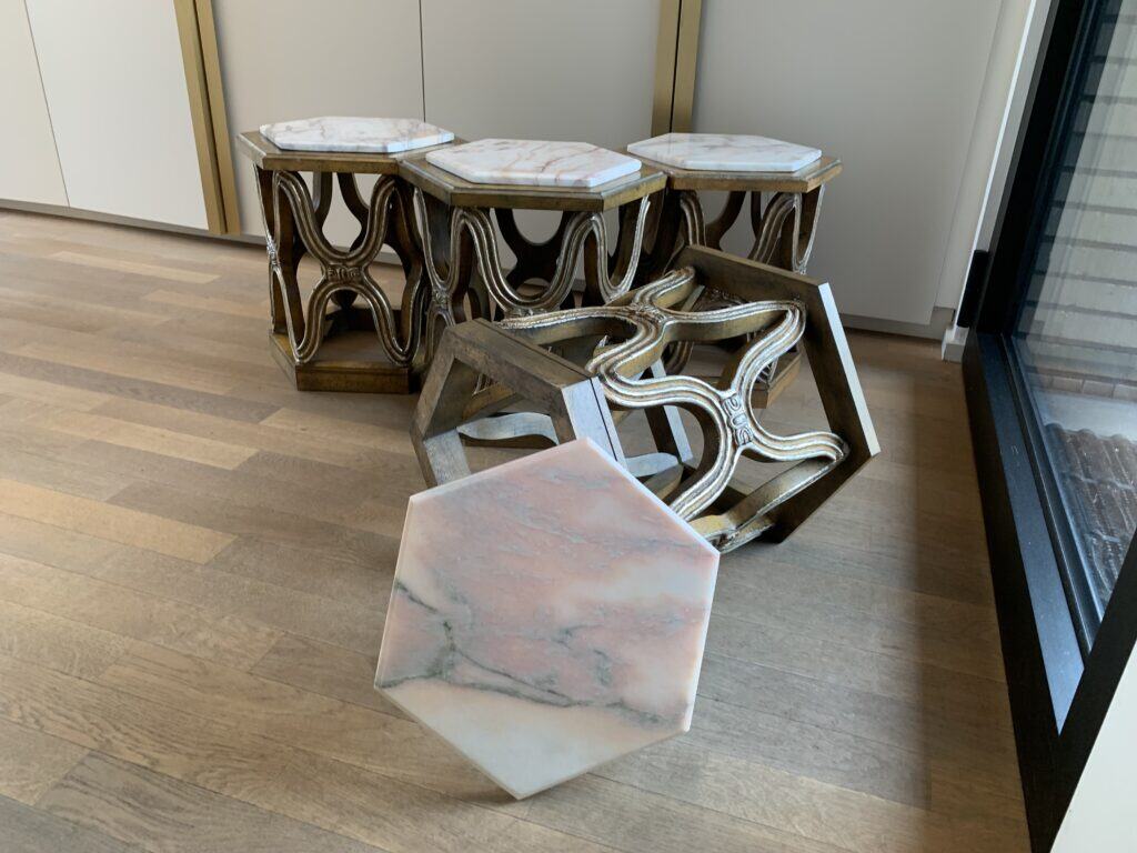 Vier sierlijke, zeshoekige bijzettafeltjes met marmeren blad staan gegroepeerd op een houten vloer. Eén tafel is omgevallen, waardoor het geometrische houten frame en de onderkant van het marmeren blad zichtbaar zijn.