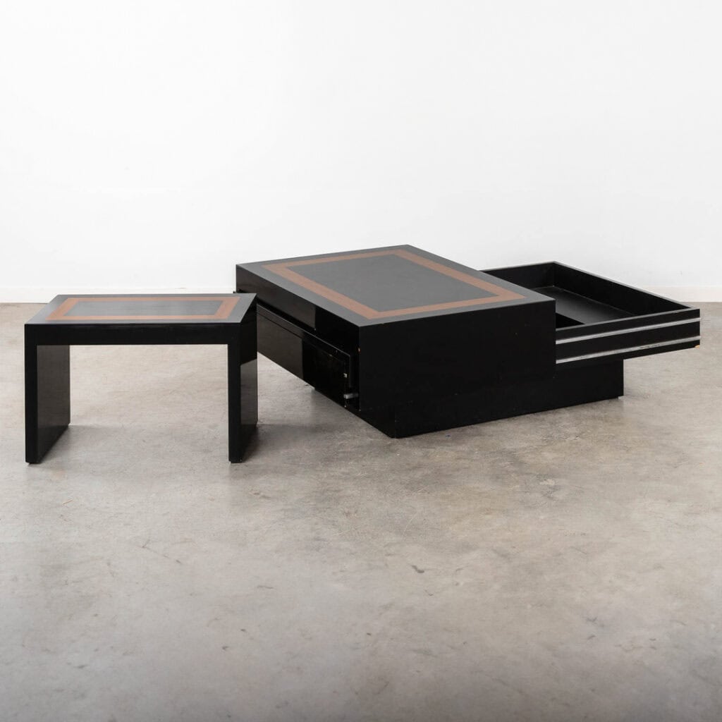 Een moderne, zwarte salontafel met houten inlegwerk, bestaande uit twee in elkaar passende tafeltjes en een uitschuifvak, tentoongesteld op een betonnen vloer tegen een witte muur.