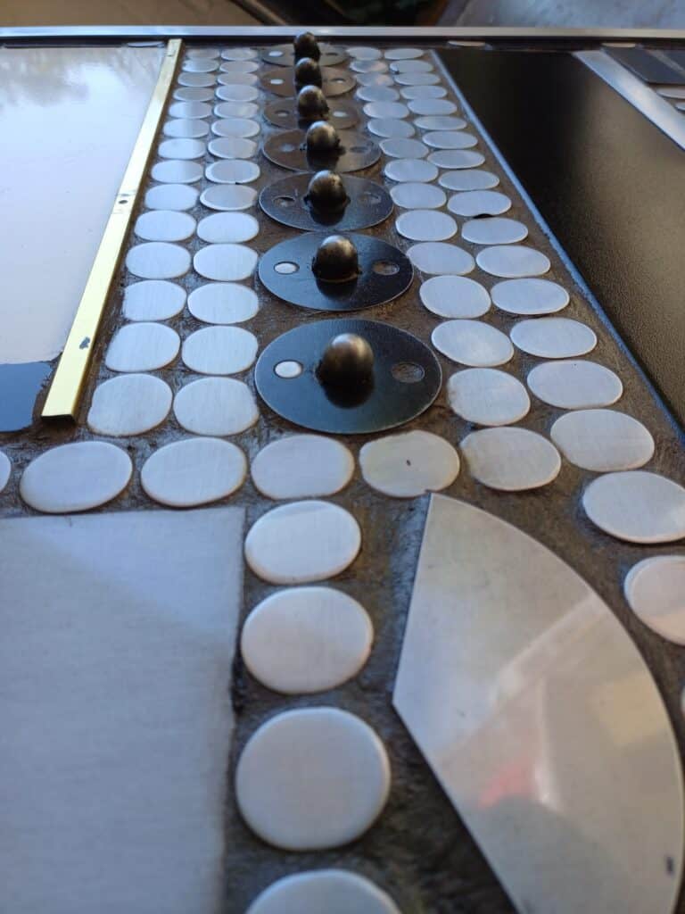 Close-up van een metalen paneel met rijen ronde zilveren schijven en zwarte bollen op ronde zwarte voetstukken, gerangschikt in een geometrisch patroon. Het oppervlak oogt industrieel en reflecterend.