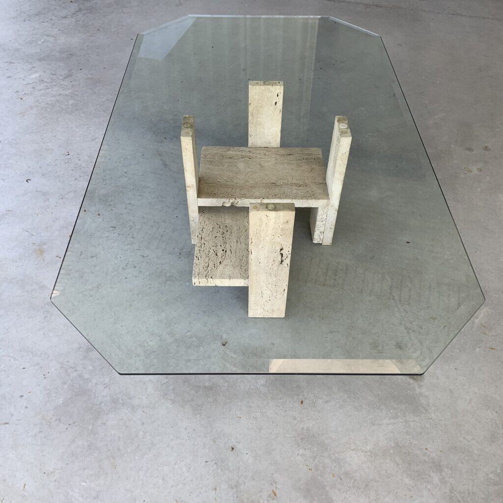 Een glazen salontafel met een achthoekig blad en een geometrisch onderstel van beige steen staat op een betonnen vloer.