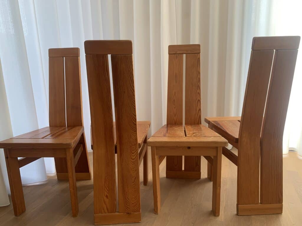Vier houten stoelen met hoge, rechte rugleuningen en een eenvoudig, robuust ontwerp staan in een groepje op een lichte houten vloer. Witte gordijnen hangen op de achtergrond en filteren het licht zachtjes.