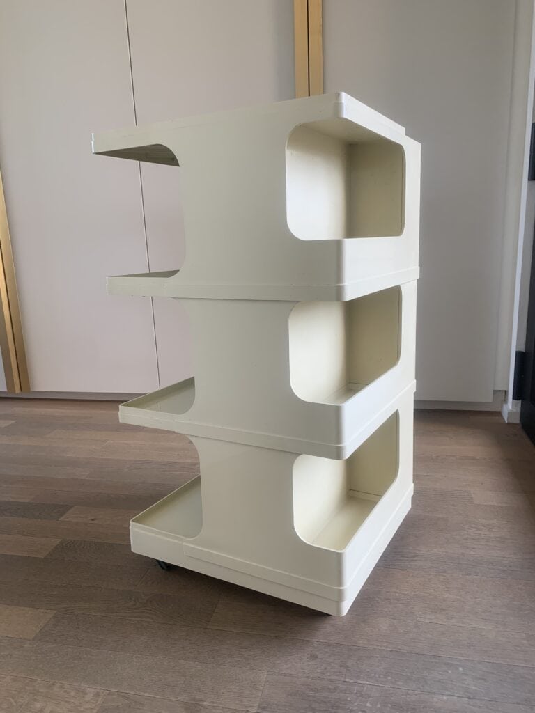Een crèmekleurige, drielaagse plastic opbergwagen met open planken en afgeronde hoeken staat op een houten vloer. De wagen staat op wielen en staat binnen bij lichtgekleurde kasten.